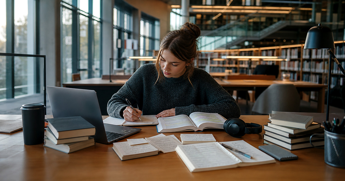 A philology student organising books, linguistic notes and research on a laptop in a library